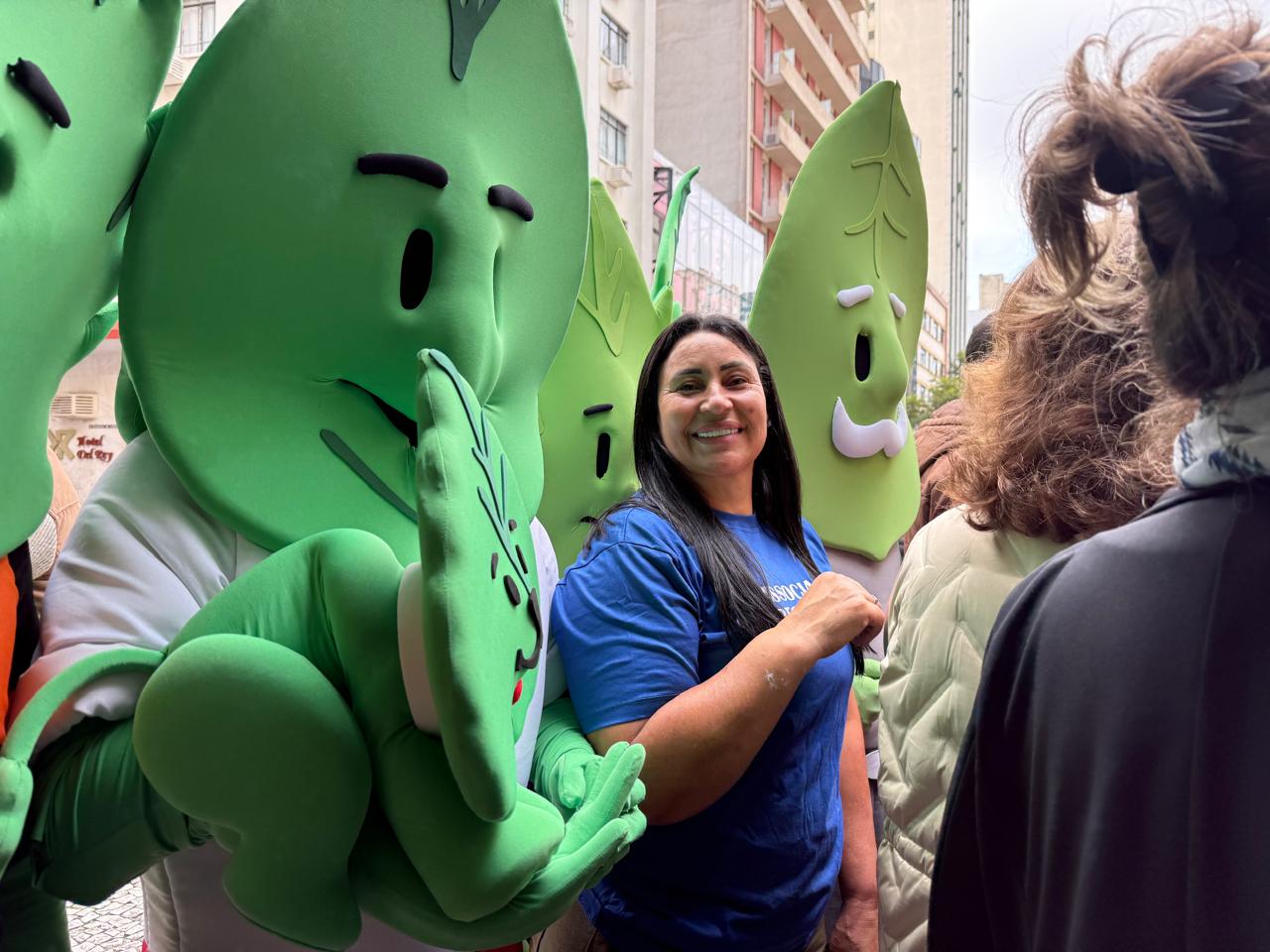 Associada da Associar Recicle Mais sorrindo ao lado dos mascotes da Família Folha durante ação de educação ambiental nas ruas de Curitiba